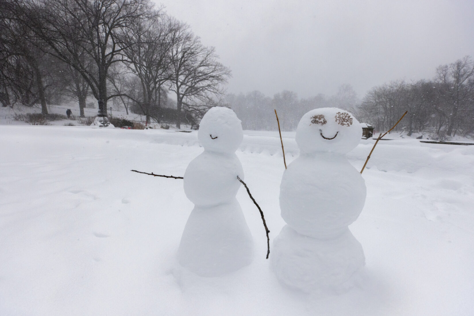 Two snowpeople smiling along the Lake in Central Park. Their arms are made of twigs and pointed up in celebration.