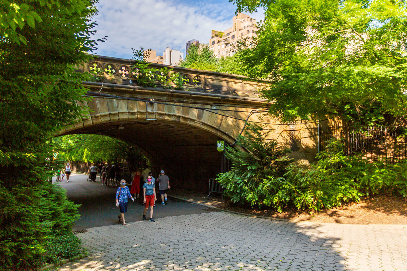 Masked parkgoers strolling under a bridge near the Children's Zoo in summer